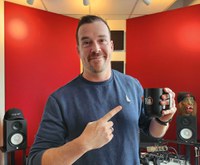 A photograph of France Brus in a recording studio wearing a hat, holding a coffee mug that says 10-year anniversary in his left hand and pointing to the mug with his right hand index finger